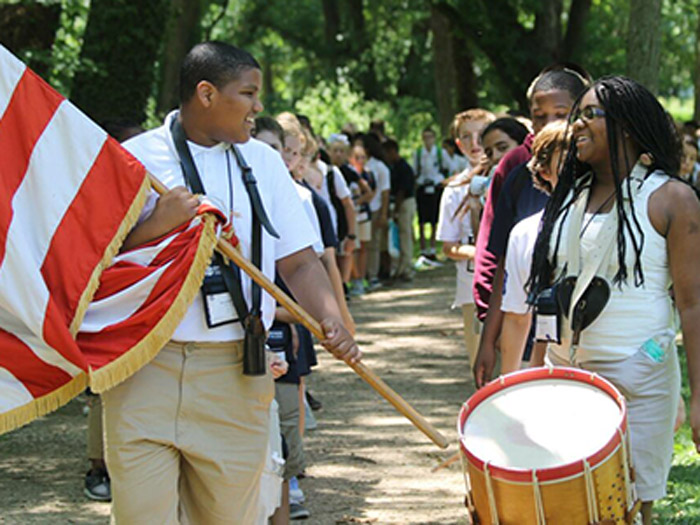 Student holding flag and camera looks over at fellow student playing drums