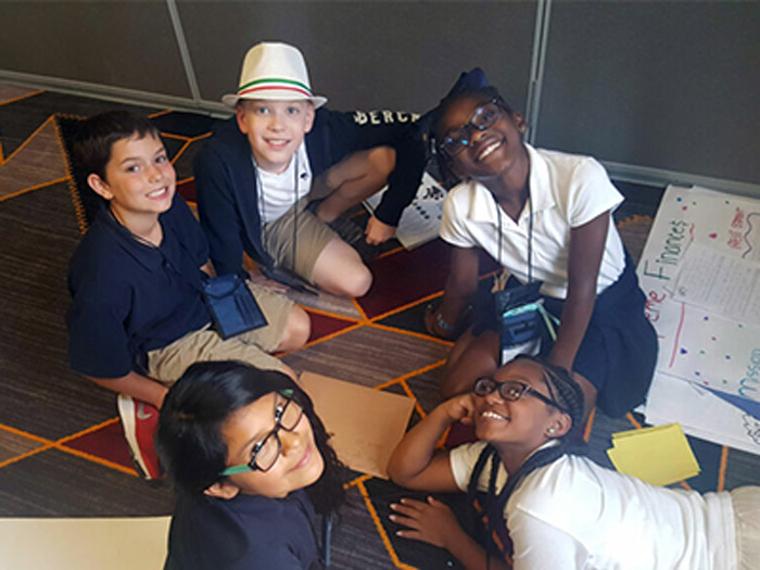 Students sitting in a circle wearing white and blue shirt with one boy wearing a white hat